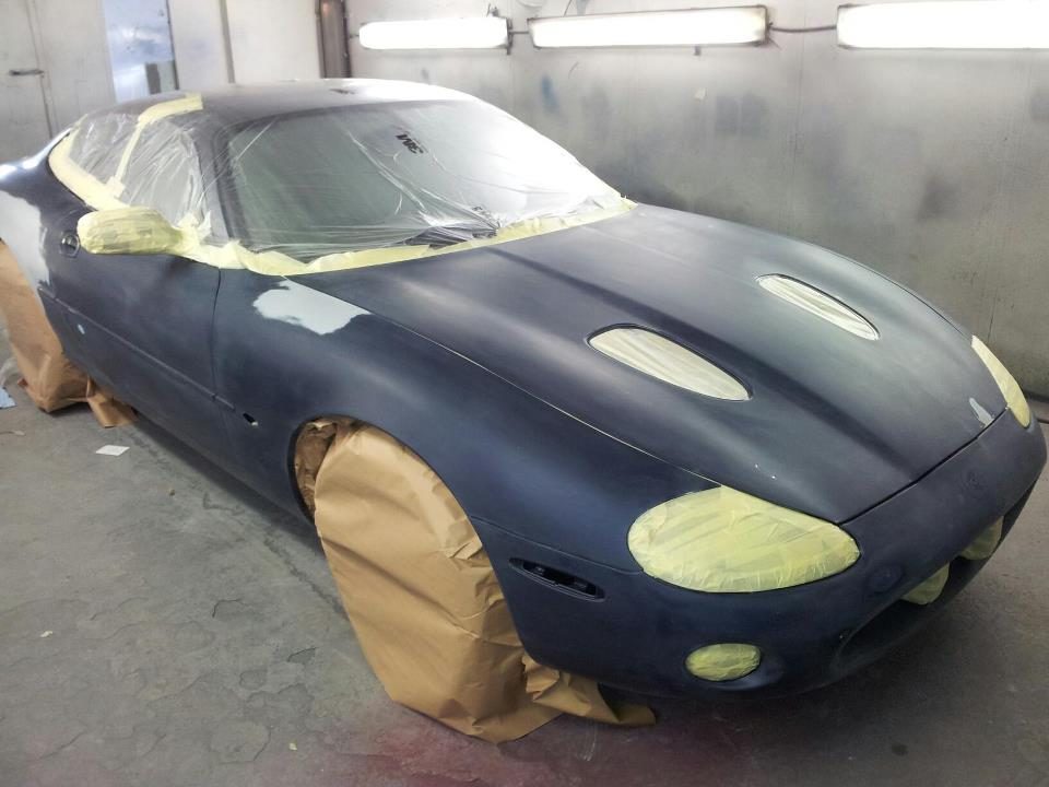 A Jaguar coupe in dark primer with lights masked in yellow tape and wheels wrapped in brown paper
